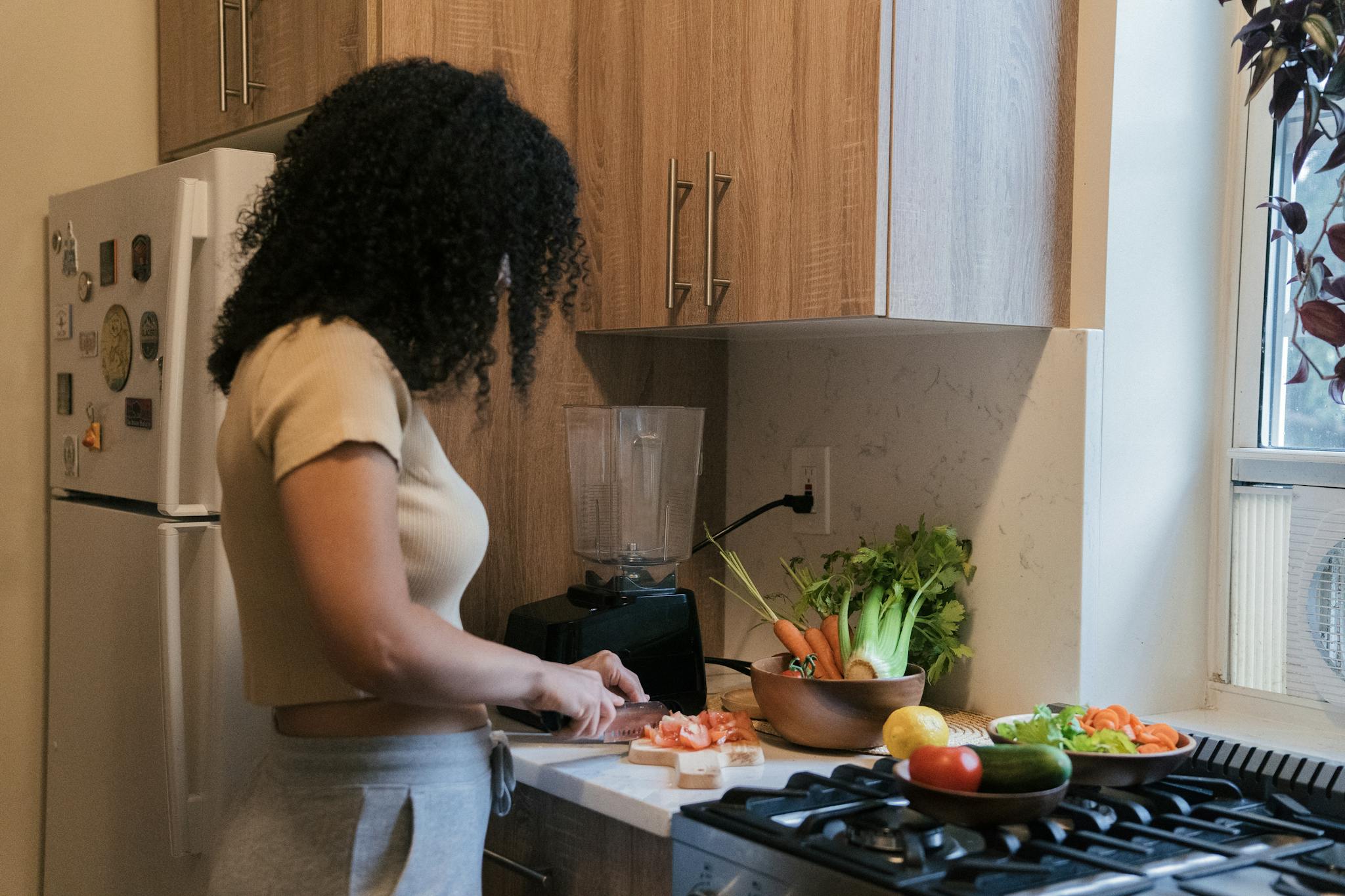 A woman cuts vegetables on a board in a modern kitchen setting, surrounded by fresh produce.