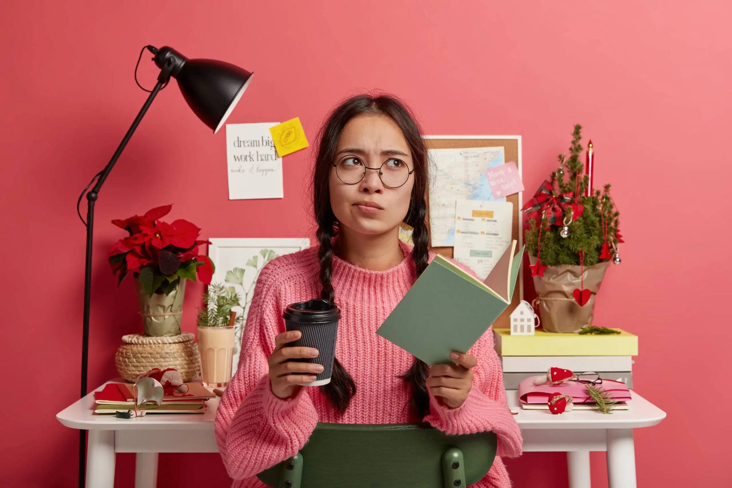Serious contemplative female wears spectacles and oversized jumper, holds paper cup of coffee, textbook for education, spends time for autodidact, ready for Christmas holiday, poses in study room